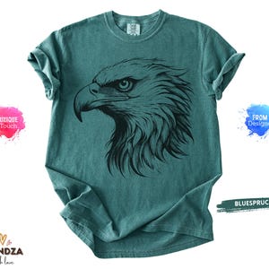 May include: Teal-colored t-shirt featuring a detailed black illustration of an eagle's head. The eagle's eye is focused, and the feathers are intricately drawn. The shirt has a classic crew neck and short sleeves. The text "BLUESPRUCE" is visible.