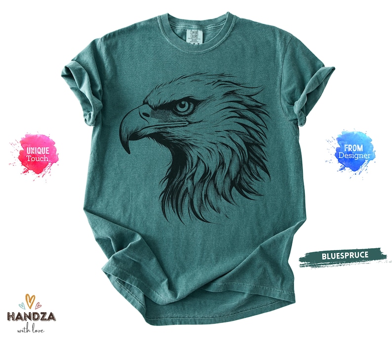 May include: Teal-colored t-shirt featuring a detailed black illustration of an eagle's head. The eagle's eye is focused, and the feathers are intricately drawn. The shirt has a classic crew neck and short sleeves. The text "BLUESPRUCE" is visible.