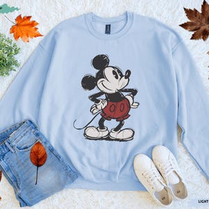 Puede incluir: Sudadera azul claro con una ilustraci&oacute;n de Mickey Mouse en negro, rojo y blanco. La imagen incluye vaqueros azules y zapatillas blancas. Las palabras "LIGHT BLUE" est&aacute;n en la parte inferior derecha. Hojas de oto&ntilde;o esparcidas.