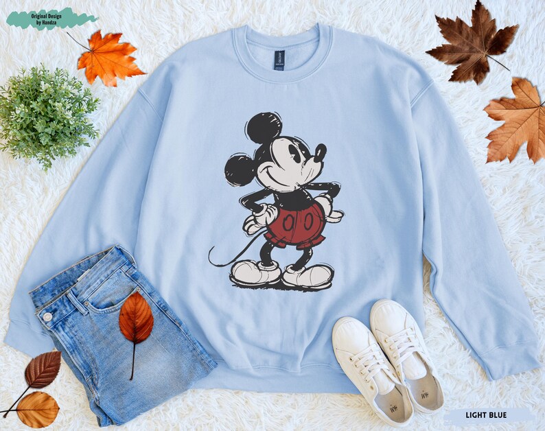 Puede incluir: Sudadera azul claro con una ilustraci&oacute;n de Mickey Mouse en negro, rojo y blanco. La imagen incluye vaqueros azules y zapatillas blancas. Las palabras "LIGHT BLUE" est&aacute;n en la parte inferior derecha. Hojas de oto&ntilde;o esparcidas.