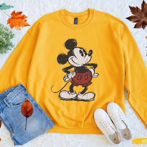 Puede incluir: Una sudadera dorada con un gr&aacute;fico de Mickey Mouse. La parte superior de manga larga tiene cuello redondo y una imagen de Mickey Mouse. La imagen se combina con unos vaqueros azules y zapatillas blancas.