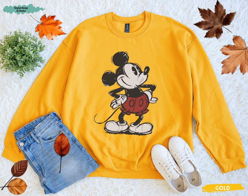 Puede incluir: Una sudadera dorada con un gr&aacute;fico de Mickey Mouse. La parte superior de manga larga tiene cuello redondo y una imagen de Mickey Mouse. La imagen se combina con unos vaqueros azules y zapatillas blancas.