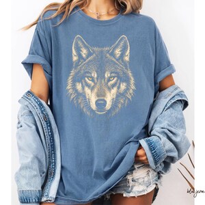 Puede incluir: Camiseta azul con una ilustraci&oacute;n detallada de la cara de un lobo en tonos dorados y marrones. Los ojos del lobo son amarillos. La camiseta forma parte de una colecci&oacute;n de ropa casual.