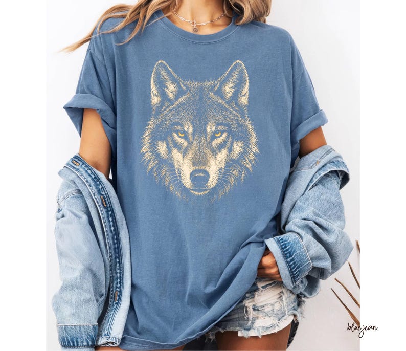 Puede incluir: Camiseta azul con una ilustraci&oacute;n detallada de la cara de un lobo en tonos dorados y marrones. Los ojos del lobo son amarillos. La camiseta forma parte de una colecci&oacute;n de ropa casual.