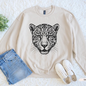 May include: A sand-colored sweatshirt featuring a black and white illustration of a leopard's face. The sweatshirt is paired with light blue jeans and white sneakers. The word "SAND" is printed in a small, light brown font.