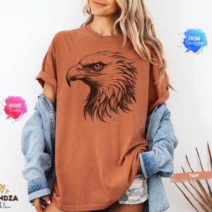 May include: A rust-colored t-shirt featuring a detailed black illustration of an eagle's head. The eagle's profile is prominently displayed, showcasing its sharp beak and intense gaze. The shirt has a relaxed fit and rolled-up sleeves.