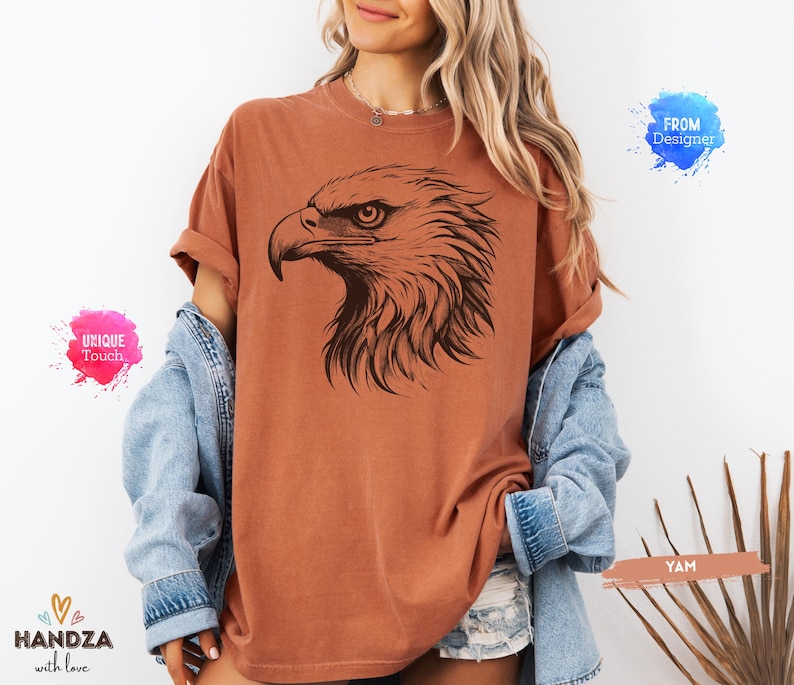 May include: A rust-colored t-shirt featuring a detailed black illustration of an eagle's head. The eagle's profile is prominently displayed, showcasing its sharp beak and intense gaze. The shirt has a relaxed fit and rolled-up sleeves.