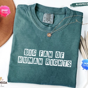 Peut inclure: Un t-shirt Comfort Colors bleu sarcelle avec le texte "BIG FAN OF HUMAN RIGHTS" en lettres blanches. Un collier doré avec un pendentif repose sur le t-shirt. Le t-shirt est plié sur une surface blanche.
