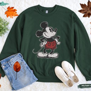 Puede incluir: Sudadera verde militar con un gr&aacute;fico desgastado de Mickey Mouse. La sudadera tiene cuello redondo y mangas largas. La imagen tambi&eacute;n muestra unos vaqueros azules y zapatillas blancas. El texto "MILITARY GREEN" es visible.