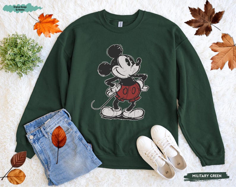 Puede incluir: Sudadera verde militar con un gr&aacute;fico desgastado de Mickey Mouse. La sudadera tiene cuello redondo y mangas largas. La imagen tambi&eacute;n muestra unos vaqueros azules y zapatillas blancas. El texto "MILITARY GREEN" es visible.