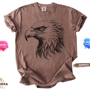 May include: Espresso-colored t-shirt featuring a detailed black illustration of an eagle's head. The design showcases the bird's sharp beak and focused gaze. The shirt has a classic crew neck and short sleeves.