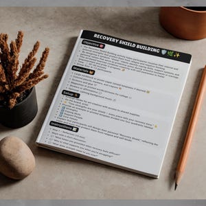 May include: A white document titled Recovery Shield Building with black text and emoji icons. The page outlines objectives, materials, setup, and instructions for an art-based activity. A wooden pencil lies beside the document on a flat surface.