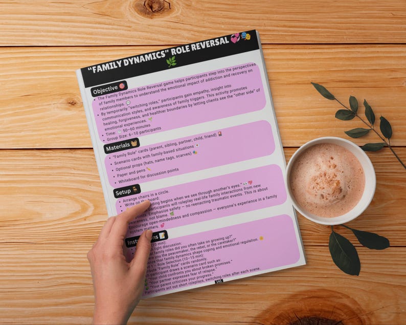 May include: A printed document titled "FAMILY DYNAMICS" ROLE REVERSAL with sections for Objective, Materials, Setup, and Instructions. The page features a light pink background with black text and various small colourful icons, presented on a wooden surface next to a white mug of coffee.