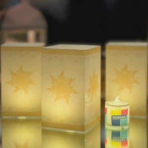 May include: Three rectangular luminaries featuring a yellow sun emblem and decorative border, illuminated from within. The text "not made of paper" is visible at the top. A small tea light candle with a "Noëlisa" label sits in the foreground.