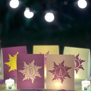 May include: A collection of rectangular table cards featuring a stylized sun emblem with the numbers 10, 5, 4, and the letter A printed in the center. The cards are purple and cream, illuminated from behind, and arranged on a surface with scattered petals.