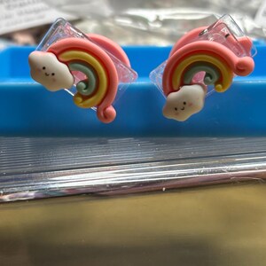 Op de afbeelding: Twee transparante plastic haarclips met regenboog- en wolkmotieven. De regenboog is veelkleurig met roze, geel, groen en blauw. De wolk is wit met een roze rand en een vrolijk gezicht.