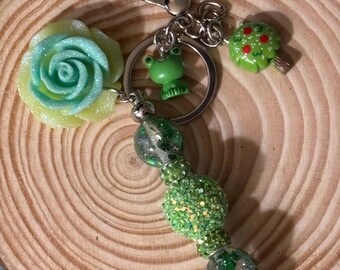 Könnte beinhalten: Grüner und silberner Schlüsselanhänger mit einem grünen Blumen-, Frosch- und Baum-Anhänger. Der Schlüsselanhänger hat auch grüne Glitzerperlen.