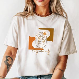 May include: Ivory-colored t-shirt featuring a minimalist design with the text "Clay vibes only." The graphic includes a sun, hands, and a vase in shades of brown and orange. The shirt is made of a soft material.