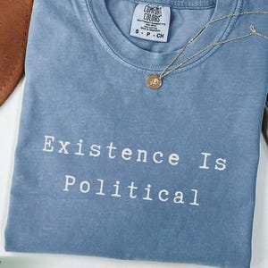 Może przedstawiać: Niebieska koszulka w kolorze dżinsu z białym napisem "Existence Is Political". Złoty naszyjnik z wisiorkiem spoczywa na koszulce. Koszulka jest złożona i obok brązowego kapelusza i suszonej rośliny.