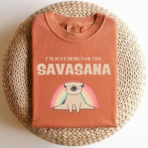 Peut inclure: T-shirt de couleur rouille avec l'inscription "I'M JUST HERE FOR THE SAVASANA" au-dessus d'un dessin animé de carlin allongé avec un arc-en-ciel. Le t-shirt est plié sur une surface circulaire tissée.