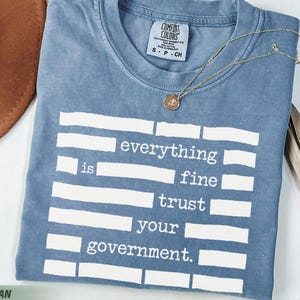 Può includere: T-shirt blu jeans Comfort Colors con la scritta bianca "everything is fine trust your government". Una collana d'oro con un ciondolo è appoggiata sulla maglietta. Un cappello marrone e una pianta secca sono sullo sfondo.