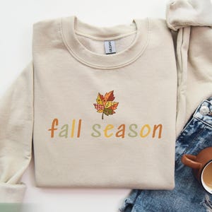 May include: A beige sweatshirt featuring the text "fall season" in autumnal colours, with a leaf design. The folded sweatshirt is next to a pair of denim jeans and a brown mug filled with a beverage.