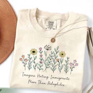 Op de afbeelding: Ivoor kleurig t-shirt met een bloemenontwerp en de tekst "Imagine Hating Immigrants More Than Pedophiles." Het shirt heeft een ronde hals en korte mouwen. Een gouden ketting met een hanger ligt op het shirt.