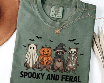 Op de afbeelding: Mosgroen t-shirt met een Halloween-thema. Het ontwerp toont vier cartoon dieren in kostuums: een spook, een pompoen, een heks en een skelet. De woorden "Spooky and Feral" staan onder de karakters gedrukt.