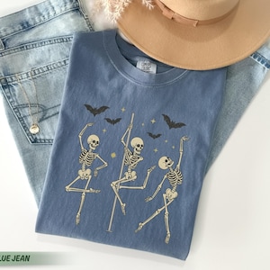 May include: A blue Comfort Colors t-shirt featuring a graphic of dancing skeletons, bats, and stars. The shirt is laid on top of light blue jeans, a gold necklace, and a tan hat. The text "BLUE JEAN" is at the bottom.