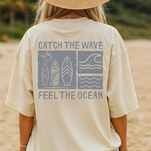 May include: Ivory-colored t-shirt with a blue graphic on the back that says "Catch the Wave, Feel the Ocean." The graphic includes a sun, surfboards, and ocean waves. The shirt is oversized and has short sleeves.