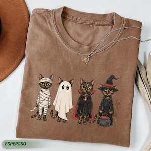 May include: A brown crew neck t-shirt featuring a graphic of four cats dressed in Halloween costumes: a mummy, a ghost, a vampire with a red-lined cape, and a witch with a black hat and a cauldron. A silver chain necklace rests on the neckline.