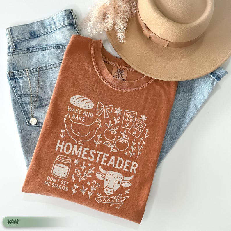 Puede incluir: Una camiseta naranja con la palabra "HOMESTEADER" y otros gr&aacute;ficos, incluyendo un pollo, pan y una vaca. La camiseta est&aacute; colocada sobre un par de vaqueros azul claro, un sombrero color canela y un collar dorado.