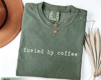 May include: A moss green t-shirt with the phrase "fueled by coffee" printed in white. A gold necklace with a pendant rests on the shirt. The shirt is folded, and a brown hat is partially visible.