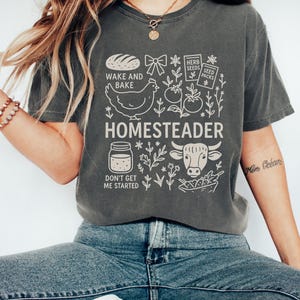 Puede incluir: Camiseta gris oscuro con un dise&ntilde;o gr&aacute;fico blanco. El dise&ntilde;o incluye la palabra "HOMESTEADER" e ilustraciones de un pollo, una vaca, pan y otros elementos relacionados con la granja. La camiseta tambi&eacute;n presenta las frases "WAKE AND BAKE" y "DON'T GET ME STARTED".