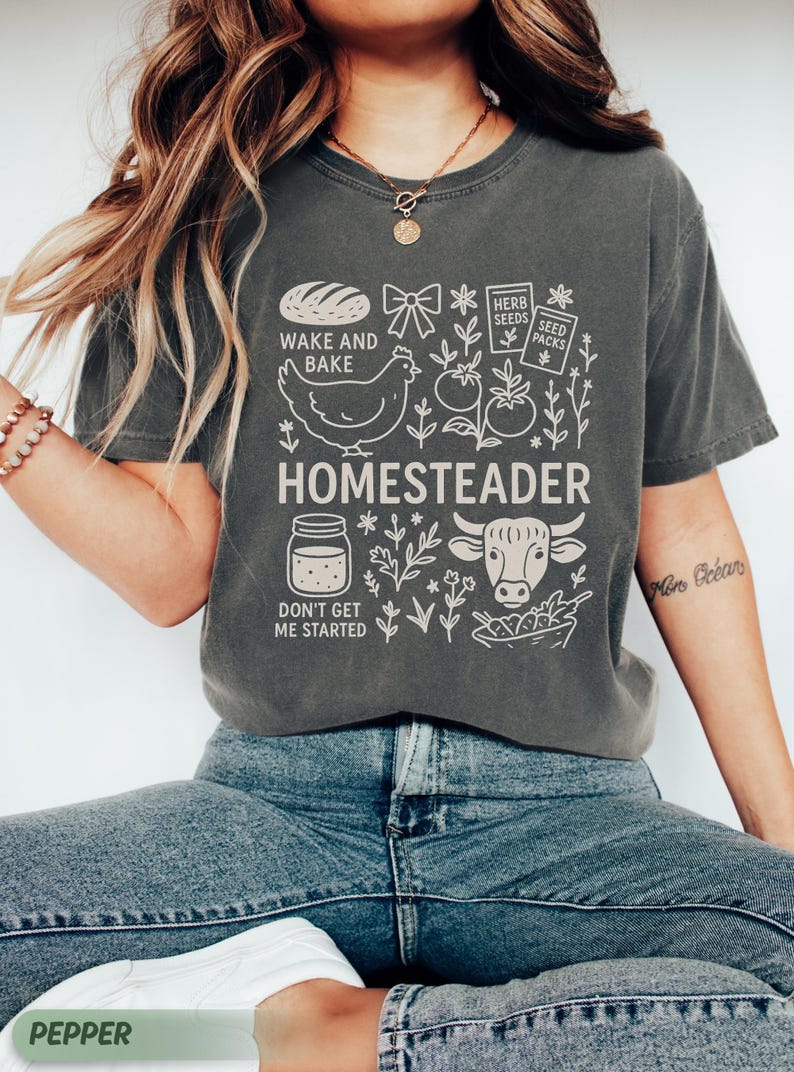 Puede incluir: Camiseta gris oscuro con un dise&ntilde;o gr&aacute;fico blanco. El dise&ntilde;o incluye la palabra "HOMESTEADER" e ilustraciones de un pollo, una vaca, pan y otros elementos relacionados con la granja. La camiseta tambi&eacute;n presenta las frases "WAKE AND BAKE" y "DON'T GET ME STARTED".