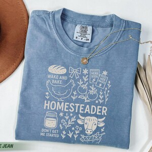 Puede incluir: Camiseta azul vaquero con un dise&ntilde;o gr&aacute;fico que presenta una gallina, pan y la palabra "HOMESTEADER". La camiseta tambi&eacute;n tiene el texto "WAKE AND BAKE" y "DON'T GET ME STARTED". Un collar de oro est&aacute; sobre la camiseta.