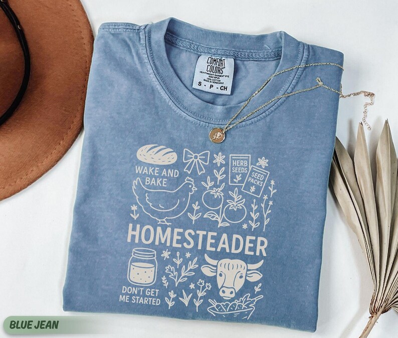 Puede incluir: Camiseta azul vaquero con un dise&ntilde;o gr&aacute;fico que presenta una gallina, pan y la palabra "HOMESTEADER". La camiseta tambi&eacute;n tiene el texto "WAKE AND BAKE" y "DON'T GET ME STARTED". Un collar de oro est&aacute; sobre la camiseta.