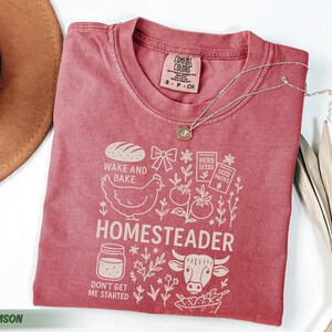 Puede incluir: Camiseta de color carmes&iacute; con un dise&ntilde;o gr&aacute;fico que presenta un pan, una gallina, tomates y la palabra "HOMESTEADER". La camiseta tambi&eacute;n incluye el texto "WAKE AND BAKE" y "DON'T GET ME STARTED".