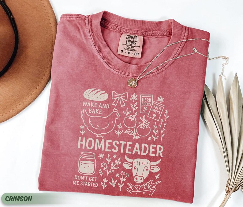 Puede incluir: Camiseta de color carmes&iacute; con un dise&ntilde;o gr&aacute;fico que presenta un pan, una gallina, tomates y la palabra "HOMESTEADER". La camiseta tambi&eacute;n incluye el texto "WAKE AND BAKE" y "DON'T GET ME STARTED".