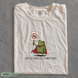 Pode incluir: Uma t-shirt cor de marfim com um sapo verde a usar uma capa vermelha e a segurar um sinal com um círculo vermelho e uma barra sobre as letras AI. Abaixo do sapo, o texto KEEP THE WEIRD LITTLE HUMAN THINGS está escrito em letras pretas.