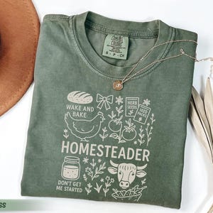 Puede incluir: Una camiseta verde musgo con un dise&ntilde;o gr&aacute;fico que presenta un pan, una gallina, tomates y la palabra "HOMESTEADER". La camiseta tambi&eacute;n incluye el texto "WAKE AND BAKE" y "DON'T GET ME STARTED."