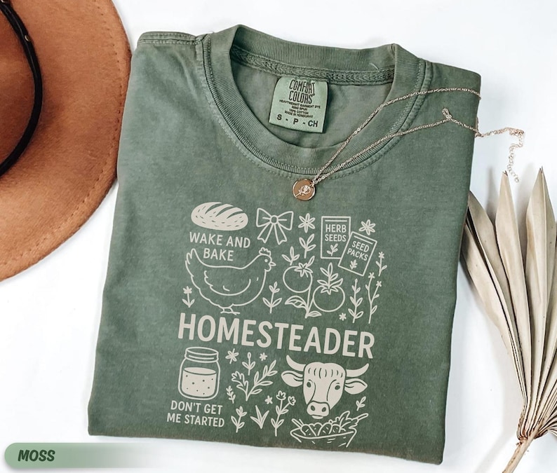 Puede incluir: Una camiseta verde musgo con un dise&ntilde;o gr&aacute;fico que presenta un pan, una gallina, tomates y la palabra "HOMESTEADER". La camiseta tambi&eacute;n incluye el texto "WAKE AND BAKE" y "DON'T GET ME STARTED."