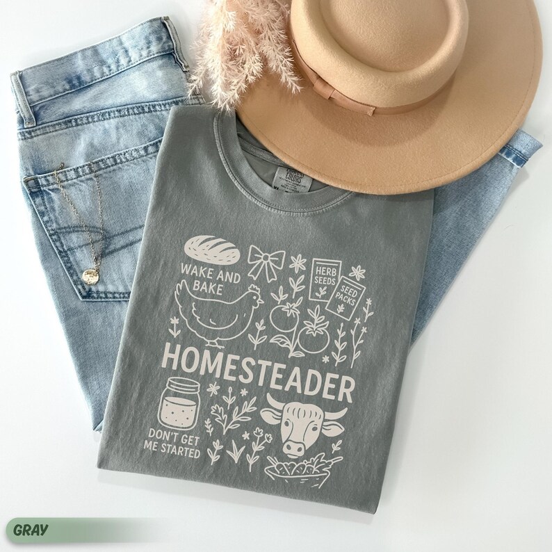 Puede incluir: Una camiseta gris con la palabra "Homestead" y gr&aacute;ficos relacionados, incluyendo un pan, una gallina, un tarro, una vaca y verduras. La camiseta se combina con vaqueros azul claro y un sombrero beige.