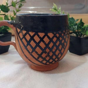 May include: A handmade ceramic mug with a brown handle and base. The mug features a black rim and a lattice design in black over a tan background. The mug is displayed with small potted plants.
