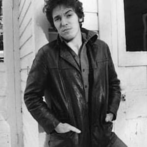May include: Black and white photograph of a person wearing a black leather jacket over a button-down shirt and jeans. The person is leaning against a white building with a window. The jacket has a collared neckline and pockets.