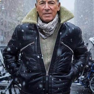 May include: A black leather jacket with a shearling collar and a gray scarf. The jacket has a zipper closure and two front pockets. The person is standing in a snowy environment.