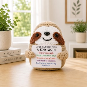 Könnte beinhalten: Gehäkelte Faultierfigur, die ein Schild mit inspirierendem Text hält. Das Faultier ist beige mit braunen Akzenten und einem weißen Gesicht. Das Schild trägt die Aufschrift "Little Reminders From A Tiny Sloth" mit positiven Affirmationen. Auf einem Holztisch.