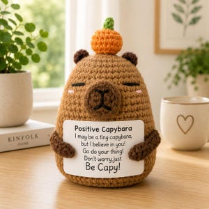 Könnte beinhalten: Gehäkelte Capybara-Figur in Brauntönen, mit einem kleinen orangefarbenen Kürbis auf dem Kopf. Das Capybara hält ein weißes Schild mit dem Text "Positive Capybara" und einer inspirierenden Botschaft. Ein charmantes, handgefertigtes Deko-Objekt.