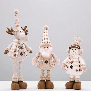 Könnte beinhalten: Drei weihnachtliche Plüschfiguren: ein Rentier, der Weihnachtsmann und ein Schneemann. Das Rentier hat braune Hörner und Stiefel, der Weihnachtsmann hat einen langen weißen Bart und der Schneemann trägt Ohrenschützer. Alle sind in Beige und Weiß mit Schneeflocken-Akzenten gekleidet.