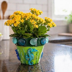 May include: A decorative, hand-painted flower pot with a vibrant yellow flowering plant. The green pot is adorned with blue seahorse and seaweed designs, and blue and clear glass beads. The pot is a charming addition to any home decor.